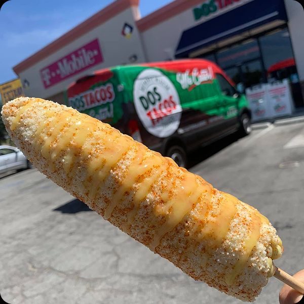 Close-up of a classic, creamy elote or Mexican street corn on a stick, drizzled with yellow sauce and chili powder, with the Dos Tacos catering van and store in the background.