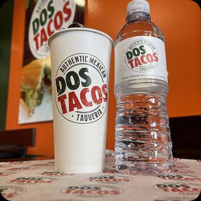 A branded paper soda cup and a plastic water bottle with the Dos Tacos Taqueria logo, sitting on a branded table liner in the restaurant, emphasizing authentic Mexican branding.