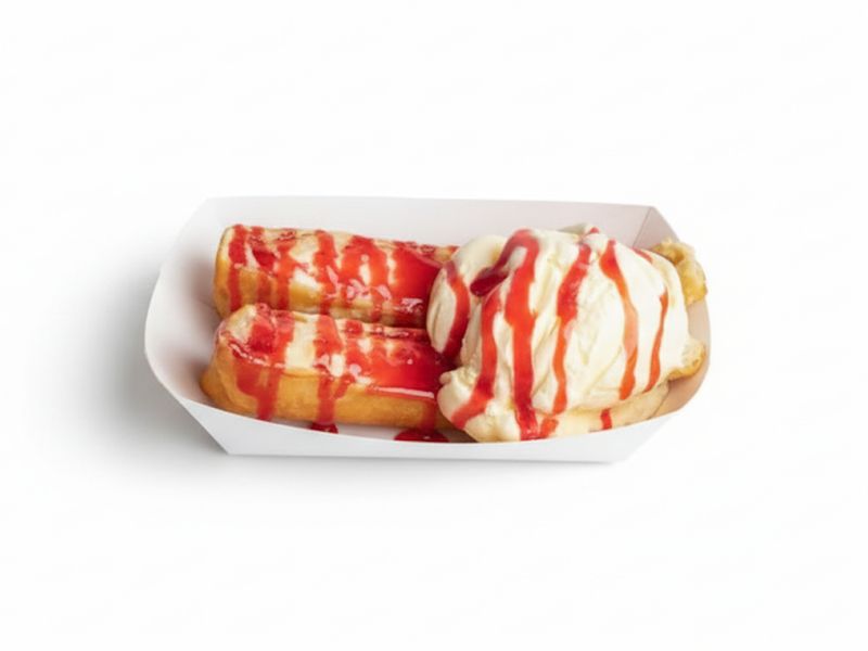 Sweet fried dough pastry dessert topped with a scoop of vanilla ice cream and drizzled with bright red strawberry syrup in a white paper tray.