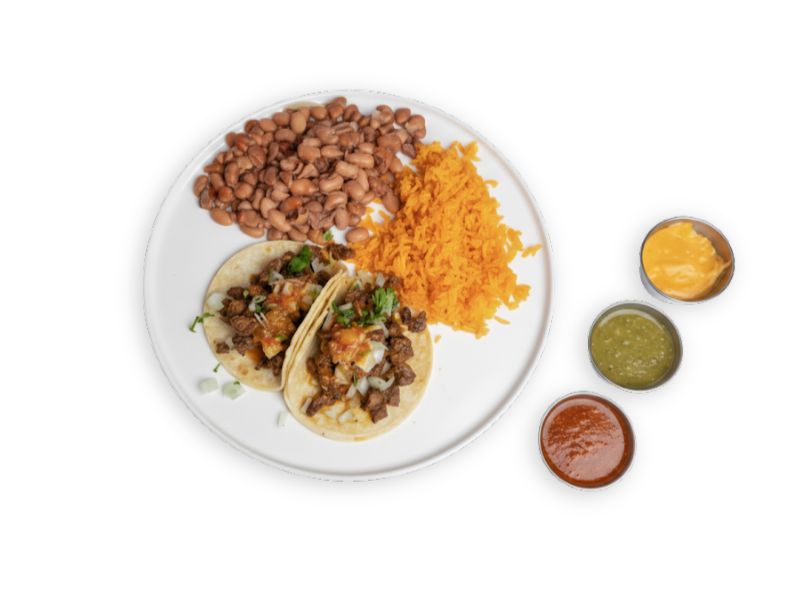 A white plate with a taco platter featuring two street tacos with carne asada and onions, a side of fluffy Mexican rice, and whole pinto beans, with three small sauce containers on the side: cheese sauce, green salsa, and red salsa.
