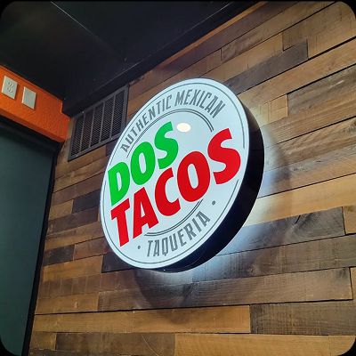 Round, illuminated sign for Dos Tacos Taqueria on an interior wood-paneled accent wall, promoting Authentic Mexican food.