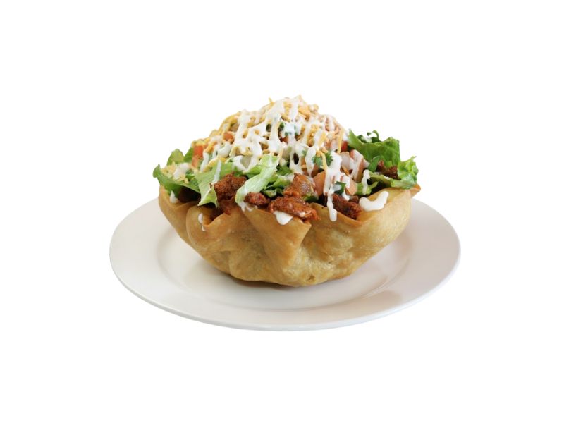 A crispy, edible flour tortilla bowl filled with a taco salad mix of seasoned meat, shredded lettuce, cheese, and sour cream on a white plate.